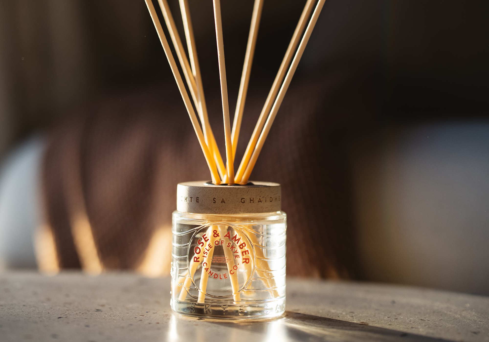 A Rose and Amber scented reed diffuser is sat on a countertop with a sofa in the background. 